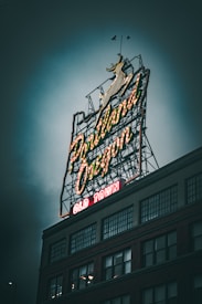 An iconic neon sign reads 'Portland Oregon' with 'Old Town' at the bottom, set against a cloudy sky. The sign is illuminated, featuring a leaping animal, likely a deer or stag, outlined in neon lights. The base of a multi-story building with large windows is visible beneath the sign, adding an urban atmosphere.