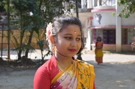 A young woman wearing traditional attire with intricate jewelry and a floral accessory in her hair. Her dress is vibrant with red and yellow colors, and she has a subtle smile. In the background, another woman in similar attire is visible, with natural greenery and a building with pink accents enhancing the scene.