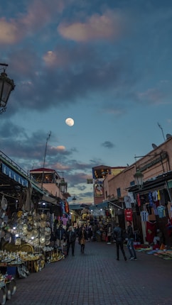 A bustling market street in the evening light, filled with various wares and lanterns hanging along the sides. The cobbled pathway is lined with stalls displaying metallic pots, clothing, and colorful textiles. The sky is a deep blue with scattered clouds and a bright full moon shining overhead.