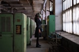 A person wearing a gas mask stands in a dimly-lit industrial setting. The scene features large green machinery and pipes on the left side and large windows on the right, casting muted light into the space.