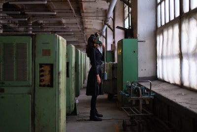 A person wearing a gas mask stands in a dimly-lit industrial setting. The scene features large green machinery and pipes on the left side and large windows on the right, casting muted light into the space.