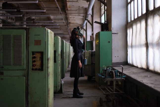 A person wearing a gas mask stands in a dimly-lit industrial setting. The scene features large green machinery and pipes on the left side and large windows on the right, casting muted light into the space.