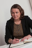 A person with shoulder-length brown hair is sitting at a table, holding a pen over an open notebook. The person is wearing a black blazer over a patterned yellow and blue top and has an expression of attentiveness.