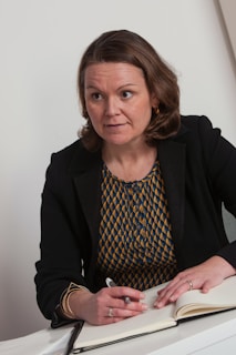 A person with shoulder-length brown hair is sitting at a table, holding a pen over an open notebook. The person is wearing a black blazer over a patterned yellow and blue top and has an expression of attentiveness.