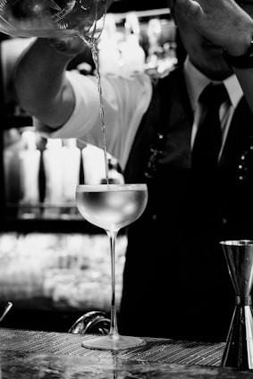 A bartender is pouring a drink from a shaker into a chilled cocktail glass. The setting appears to be a dimly lit bar with blurred lights in the background. The bartender is wearing a vest and tie, and the focus is on the elegance of the pouring action.