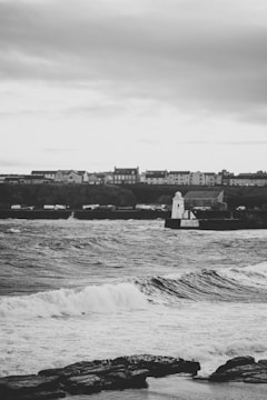 A coastal scene featuring a row of houses atop a rocky shoreline. Rough waves crash against the shore, with a lighthouse situated on a pier extending into the sea. The sky is overcast, adding a dramatic effect to the scene.