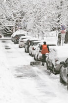 A snow-covered street with parked cars lined along the side, each coated in a thick layer of snow. Branches of trees hang overhead, heavily laden with snow. A person in an orange vest shovels snow from the sidewalk, indicating an effort to clear the path. Nearby, a road sign is visible, also covered in snow.