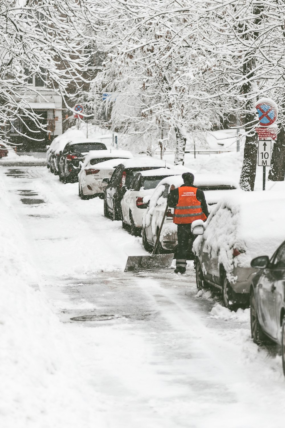 man wearing orange vest holding black snow shovel on snow street near the cars during winter