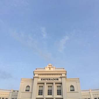 An elegant white building with classical architectural features, adorned with columns and decorative elements, topped with signage that reads 'EMPERADOR'. The sky is clear and blue, providing a serene backdrop.