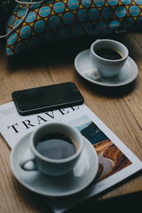 Two cups of coffee sit on saucers atop a wooden table, along with a travel magazine and a black smartphone. A colorful cushion with a blue and brown geometric pattern is visible in the background.