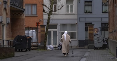 An elderly individual wearing a long beige coat and a white headscarf walks down an urban alleyway using a walking cane. Surrounding structures consist of brick and stucco buildings, with visible windows, doors, and some trash bins. A leafless tree is present in the middle of the alley. The general setting appears relatively quiet and somewhat deserted.