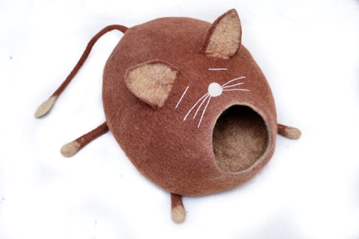 A felted wool cat bed designed to resemble a stylized orange cat with pointy ears, whiskers, and a tail. The entrance to the bed is a circular hole where the cat's mouth would be. The cat bed is mostly orange with beige accents for the ears and tail.