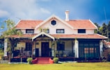 A charming house with a vintage design featuring a red tiled roof and white walls. The building has a symmetrical facade with a central entrance. The front porch is covered with vines and surrounded by a well-maintained lawn.