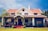 A charming house with a vintage design featuring a red tiled roof and white walls. The building has a symmetrical facade with a central entrance. The front porch is covered with vines and surrounded by a well-maintained lawn.