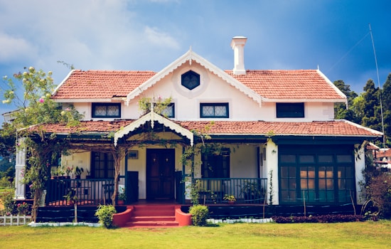A charming house with a vintage design featuring a red tiled roof and white walls. The building has a symmetrical facade with a central entrance. The front porch is covered with vines and surrounded by a well-maintained lawn.