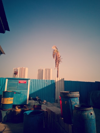 An urban construction site features a variety of industrial elements such as a generator, large barrels, and building materials scattered around. In the background, high-rise buildings are partially visible behind a blue fence, and a lone palm tree stands nearby swaying gently.