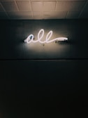 A glowing neon sign displaying the word 'all' is mounted on a textured dark wall, casting a soft light that highlights the surrounding surface patterns. The ceiling above is tiled.