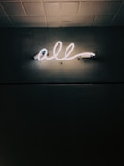 A glowing neon sign displaying the word 'all' is mounted on a textured dark wall, casting a soft light that highlights the surrounding surface patterns. The ceiling above is tiled.