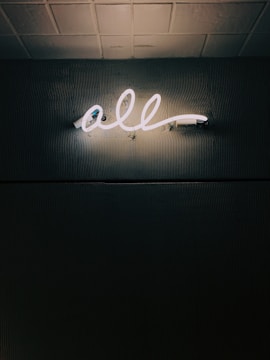 A glowing neon sign displaying the word 'all' is mounted on a textured dark wall, casting a soft light that highlights the surrounding surface patterns. The ceiling above is tiled.