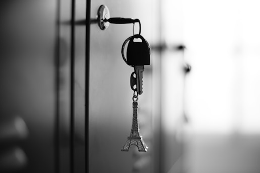 A set of keys is inserted into a lock on a smooth surface, with an Eiffel Tower keychain hanging from the keys. The focus is on the keys and the lock, while the background is softly blurred, creating a shallow depth of field effect. The image is in black and white, emphasizing contrast and light.