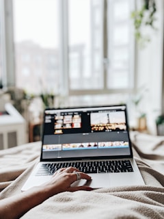 A person is lying in bed with a laptop on their legs, working on video editing software. The room is softly lit with natural light coming through large windows, and there are plants visible in the background.