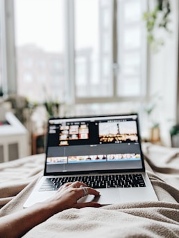 A person is lying in bed with a laptop on their legs, working on video editing software. The room is softly lit with natural light coming through large windows, and there are plants visible in the background.