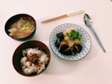 A Japanese meal consisting of a bowl of miso soup with tofu and green onions, a bowl of white rice topped with seasoning, and a dish featuring root vegetables and mushrooms garnished with fresh cilantro. The meal is set on a white table accompanied by chopsticks and a spoon.