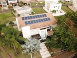 A modern house features solar panels on the roof, surrounded by lush greenery including palm trees. The property is part of a suburban neighborhood with well-maintained lawns and neighboring houses. Paved walkways lead to the main entrance, and a tree casts shade on the driveway.