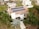 A modern house features solar panels on the roof, surrounded by lush greenery including palm trees. The property is part of a suburban neighborhood with well-maintained lawns and neighboring houses. Paved walkways lead to the main entrance, and a tree casts shade on the driveway.