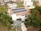 A modern house features solar panels on the roof, surrounded by lush greenery including palm trees. The property is part of a suburban neighborhood with well-maintained lawns and neighboring houses. Paved walkways lead to the main entrance, and a tree casts shade on the driveway.