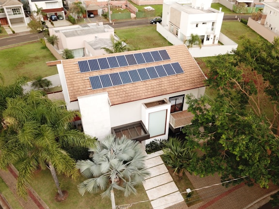 A modern house features solar panels on the roof, surrounded by lush greenery including palm trees. The property is part of a suburban neighborhood with well-maintained lawns and neighboring houses. Paved walkways lead to the main entrance, and a tree casts shade on the driveway.
