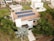 A modern house features solar panels on the roof, surrounded by lush greenery including palm trees. The property is part of a suburban neighborhood with well-maintained lawns and neighboring houses. Paved walkways lead to the main entrance, and a tree casts shade on the driveway.