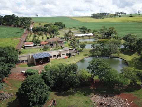 An aerial view of a rural landscape featuring a farmhouse surrounded by lush greenery and multiple small ponds. The property includes solar panels on the building roofs, and is set amidst rolling fields and scattered trees. The sky is overcast with clouds, adding depth to the scenery.