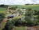An aerial view of a rural landscape featuring a farmhouse surrounded by lush greenery and multiple small ponds. The property includes solar panels on the building roofs, and is set amidst rolling fields and scattered trees. The sky is overcast with clouds, adding depth to the scenery.