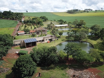 An aerial view of a rural landscape featuring a farmhouse surrounded by lush greenery and multiple small ponds. The property includes solar panels on the building roofs, and is set amidst rolling fields and scattered trees. The sky is overcast with clouds, adding depth to the scenery.