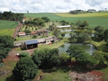 An aerial view of a rural landscape featuring a farmhouse surrounded by lush greenery and multiple small ponds. The property includes solar panels on the building roofs, and is set amidst rolling fields and scattered trees. The sky is overcast with clouds, adding depth to the scenery.