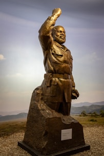 A bronze statue of a bearded man in military attire with his right arm raised in a salute or gesture of strength. The figure stands on a rocky base with a plaque attached. The background features a natural landscape with grassy plains and distant hills under a blue sky.