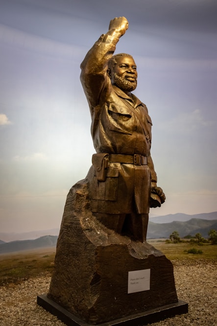 A bronze statue of a bearded man in military attire with his right arm raised in a salute or gesture of strength. The figure stands on a rocky base with a plaque attached. The background features a natural landscape with grassy plains and distant hills under a blue sky.