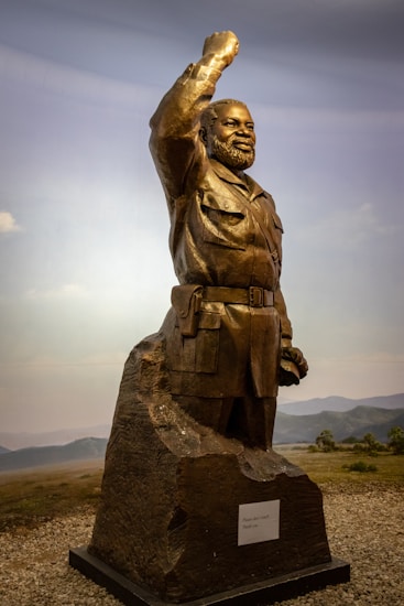 A bronze statue of a bearded man in military attire with his right arm raised in a salute or gesture of strength. The figure stands on a rocky base with a plaque attached. The background features a natural landscape with grassy plains and distant hills under a blue sky.