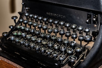 A close-up view of an old-fashioned typewriter, highlighting the round black keys with white letters. The keys appear slightly worn, showing signs of age and use. The machine itself has a rugged, vintage appearance, with some dust collecting around the base.