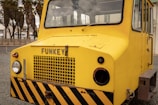 A bright yellow industrial vehicle with a flat front and large windows is situated outdoors. The vehicle has the word 'FUNKEY' displayed on its front and features black and yellow warning stripes at the base. Visible beside the vehicle are some palm trees and a building structure in the background.
