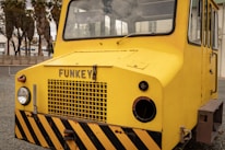 A bright yellow industrial vehicle with a flat front and large windows is situated outdoors. The vehicle has the word 'FUNKEY' displayed on its front and features black and yellow warning stripes at the base. Visible beside the vehicle are some palm trees and a building structure in the background.