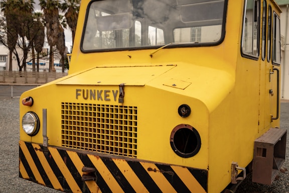 A bright yellow industrial vehicle with a flat front and large windows is situated outdoors. The vehicle has the word 'FUNKEY' displayed on its front and features black and yellow warning stripes at the base. Visible beside the vehicle are some palm trees and a building structure in the background.