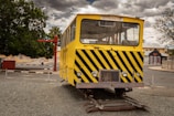 A yellow rail inspection vehicle with black stripes is positioned on short, curved tracks in an outdoor setting. The vehicle is surrounded by a quiet urban landscape with cloudy skies, a tree, and distant buildings.