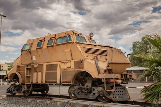 A large, heavily armored vehicle with a robust, industrial design rests on a piece of railway track. The vehicle features a tan, rusty exterior with metal plating and multiple windows. Its shape is reminiscent of a train wagon but appears fortified for heavy-duty use. The background shows a cloudy sky with some patches of blue, along with nearby trees and parts of a building.