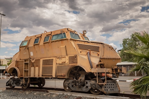 A large, heavily armored vehicle with a robust, industrial design rests on a piece of railway track. The vehicle features a tan, rusty exterior with metal plating and multiple windows. Its shape is reminiscent of a train wagon but appears fortified for heavy-duty use. The background shows a cloudy sky with some patches of blue, along with nearby trees and parts of a building.