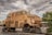 A large, heavily armored vehicle with a robust, industrial design rests on a piece of railway track. The vehicle features a tan, rusty exterior with metal plating and multiple windows. Its shape is reminiscent of a train wagon but appears fortified for heavy-duty use. The background shows a cloudy sky with some patches of blue, along with nearby trees and parts of a building.