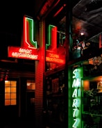 A nighttime street scene featuring neon signs with the words 'Magic Mushrooms' in red and 'Smartz' in green. The signs reflect in a window, providing a vibrant glow against the darkness. The surrounding area includes windows with soft yellow lighting, adding depth to the urban setting.