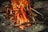 A campfire with vivid flames rises among neatly arranged rocks. The firewood is comprised of sticks and branches, generating a warm, glowing light.