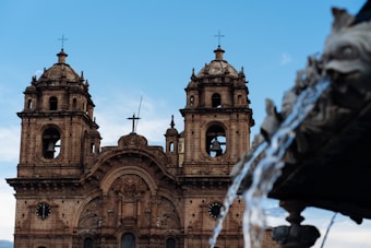 A historical church with two bell towers stands prominently against a clear blue sky. Ornate architectural details adorn the facade, and a cross is visible atop the central structure. In the foreground, water flows gracefully from a fountain decorated with sculptural elements.